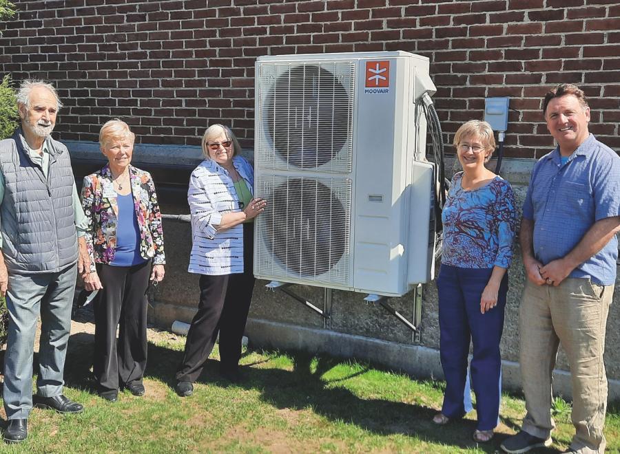 Five people stand in front of a building next to a heat pump. The heat pump is in the middle. Starting from the left is an older white man with grey hair. He is wearing a button up shirt and jeans with a navy vest. Next to him is an older woman with short white hair in a blue shirt and a floral blazer and black pants. Next to her is an older white woman with white hair, brown sunglasses and a striped white and black shirt with black pants. Her right hand rests on the white heat pump, which has two vertical fans. Next to the heat pump is an older white woman with white hair, glasses, a floral top and black pants. Next to her is a middle-aged white man with brown hair, blue shirt and beige slacks.