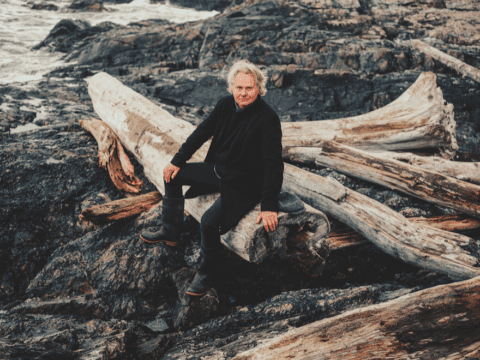 A middle-aged white man sits on logs by the beach. He has white hair and is wearing all black.