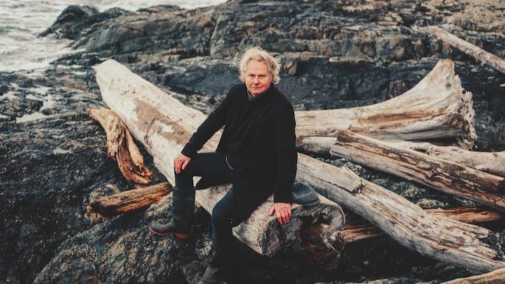 A middle-aged white man sits on logs by the beach. He has white hair and is wearing all black.