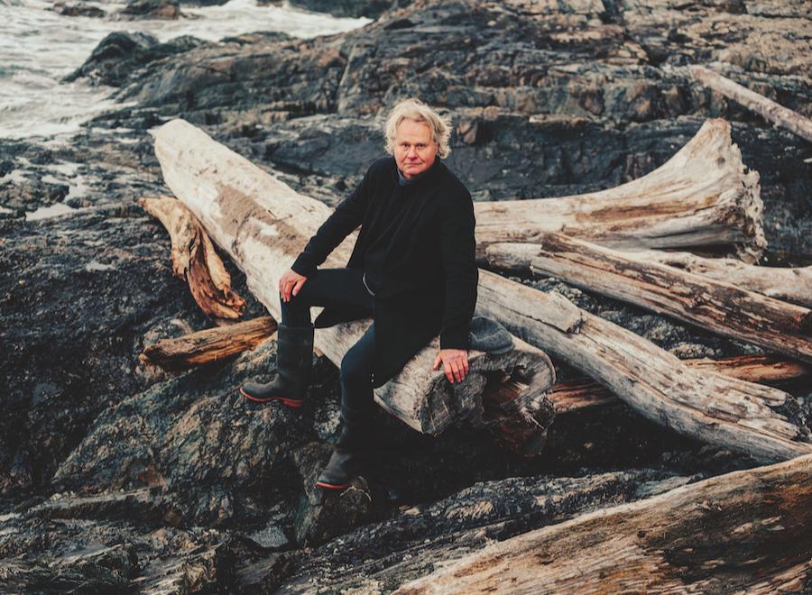 A middle-aged white man sits on logs by the beach. He has white hair and is wearing all black.