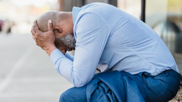 A black man wearing a blue shirt and jeans is sitting with his head in his hands.