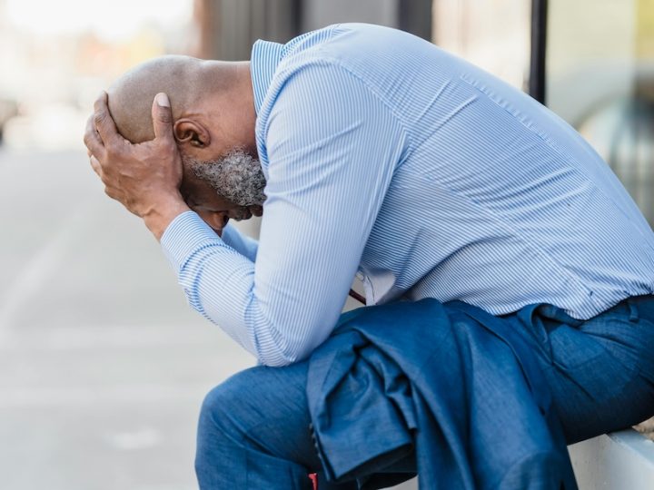 A black man wearing a blue shirt and jeans is sitting with his head in his hands.