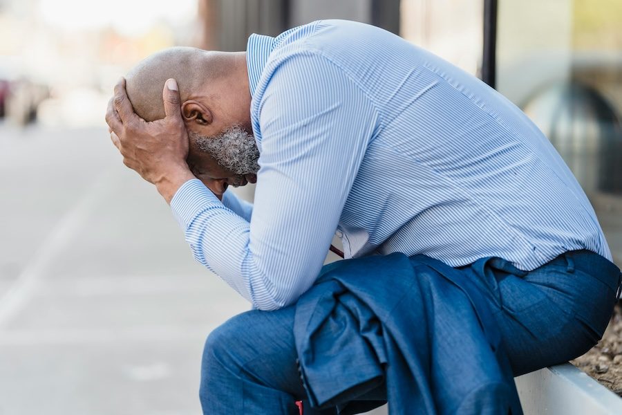 A black man wearing a blue shirt and jeans is sitting with his head in his hands.