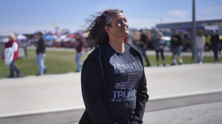 A white woman with curly grey hair poses. She is wearing a navy jacket with a t-shirt that says, "Trump is my saviour."