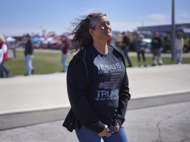 A white woman with curly grey hair poses. She is wearing a navy jacket with a t-shirt that says, "Trump is my saviour."