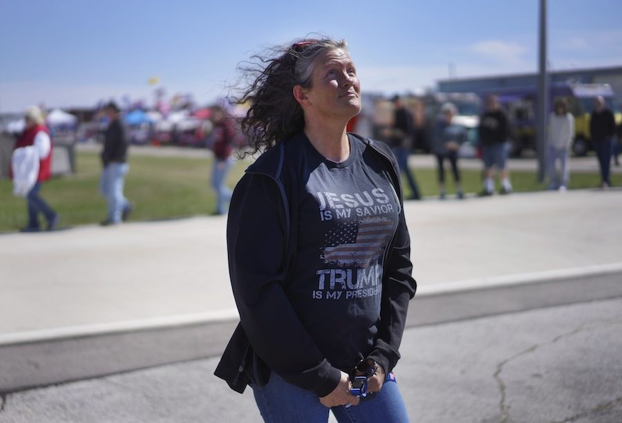 A white woman with curly grey hair poses. She is wearing a navy jacket with a t-shirt that says, "Trump is my saviour."