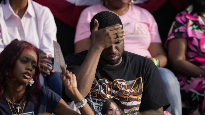 A Black man wearing a navy shirt with the American flag has his head in his hands. A Black woman in a navy t-shirt sits next to him and they are in a crowd.