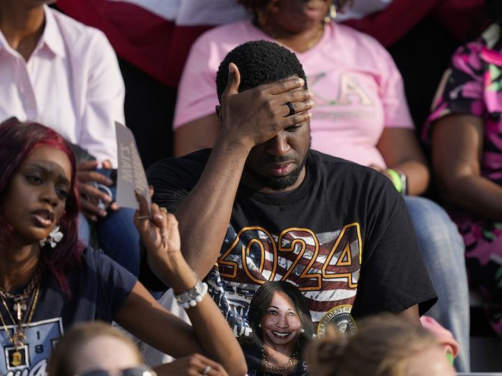 A Black man wearing a navy shirt with the American flag has his head in his hands. A Black woman in a navy t-shirt sits next to him and they are in a crowd.