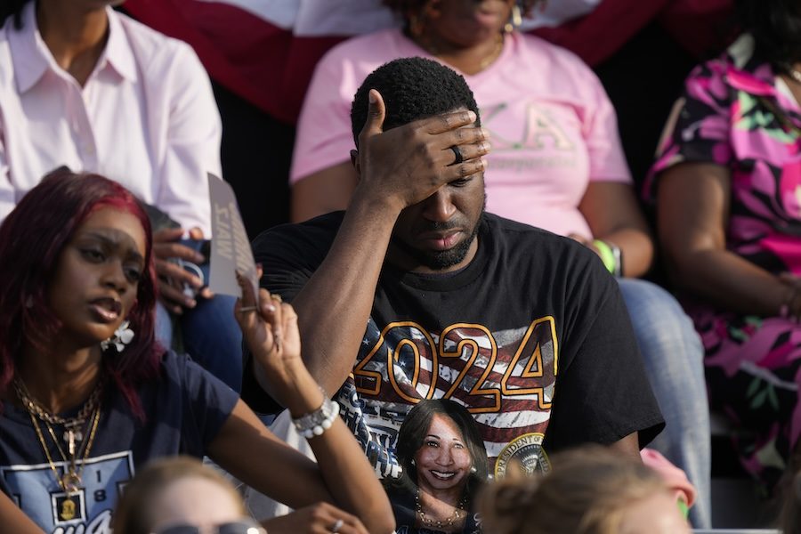 A Black man wearing a navy shirt with the American flag has his head in his hands. A Black woman in a navy t-shirt sits next to him and they are in a crowd.