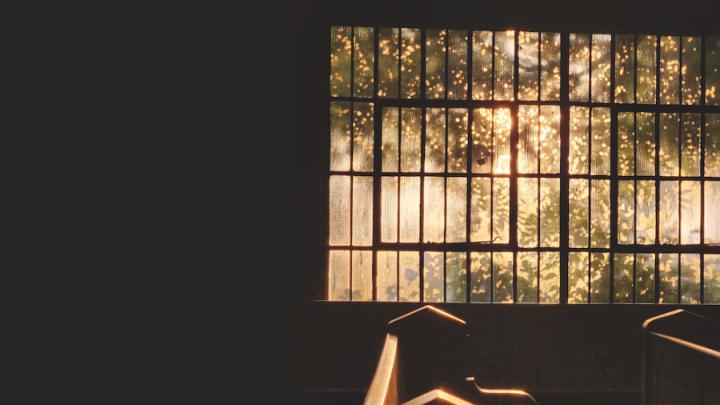 The sun shines through large panes of glass in a window and pews are visible.