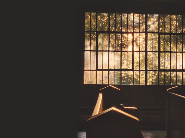 The sun shines through large panes of glass in a window and pews are visible.