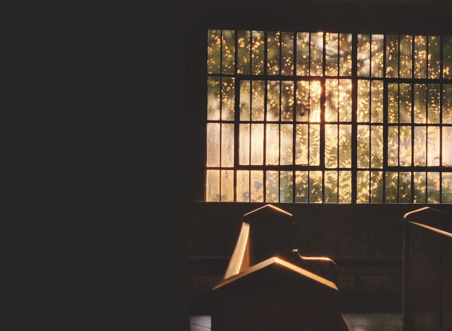 The sun shines through large panes of glass in a window and pews are visible.