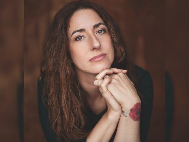 Headshot of a white woman. She has brown hair, brown eyes and a red flower tattoo on her left wrist.