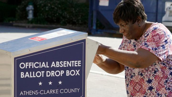 A Black woman in a dress places a ballot in a box labelled "official absentee ballot drop box"