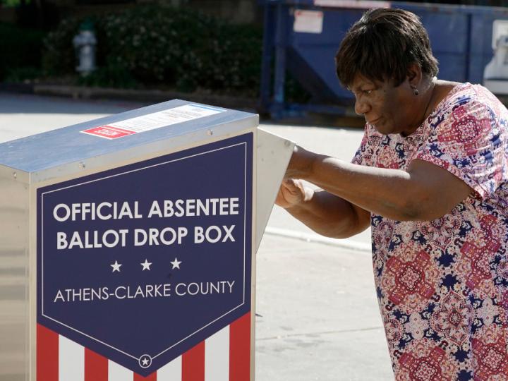 A Black woman in a dress places a ballot in a box labelled "official absentee ballot drop box"