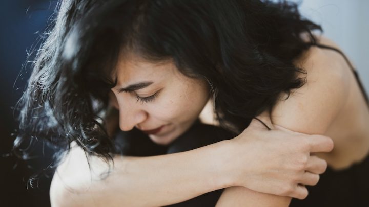 A white woman with dark brown hair sits with her head lowered and she is holding her arms