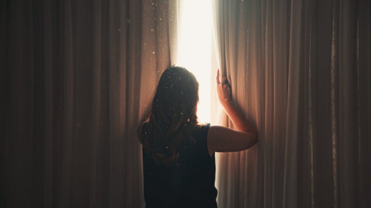 woman stands in front of curtain and opens curtains to let light in