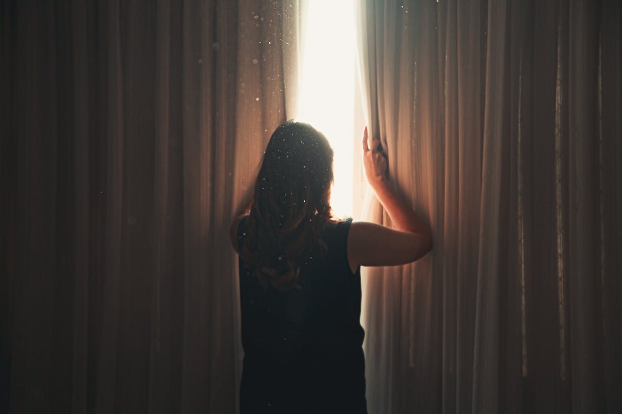 woman stands in front of curtain and opens curtains to let light in