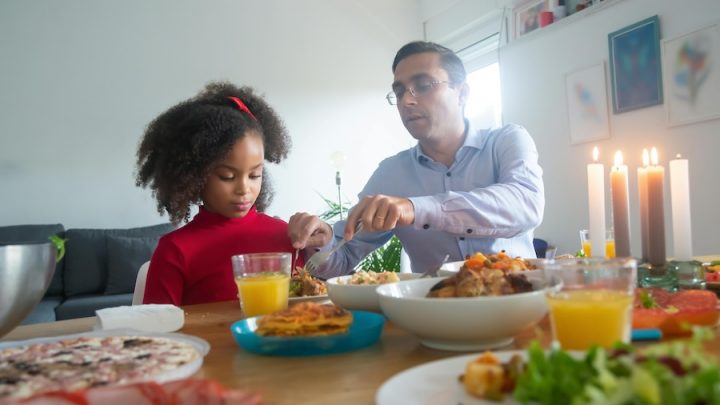 A white man in a button up shirt and glasses serves food to a Black girl with curly black hair, a red bow and red shirt. On the table there is a bowl of food, a glass of orange juice and a plate.