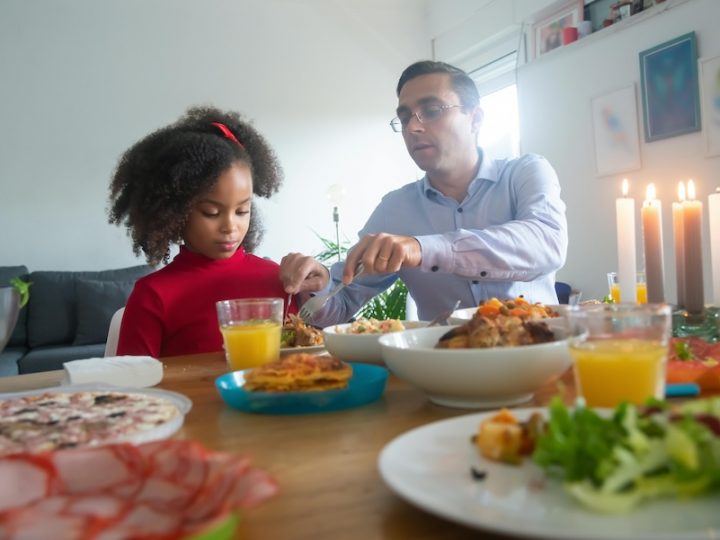 A white man in a button up shirt and glasses serves food to a Black girl with curly black hair, a red bow and red shirt. On the table there is a bowl of food, a glass of orange juice and a plate.