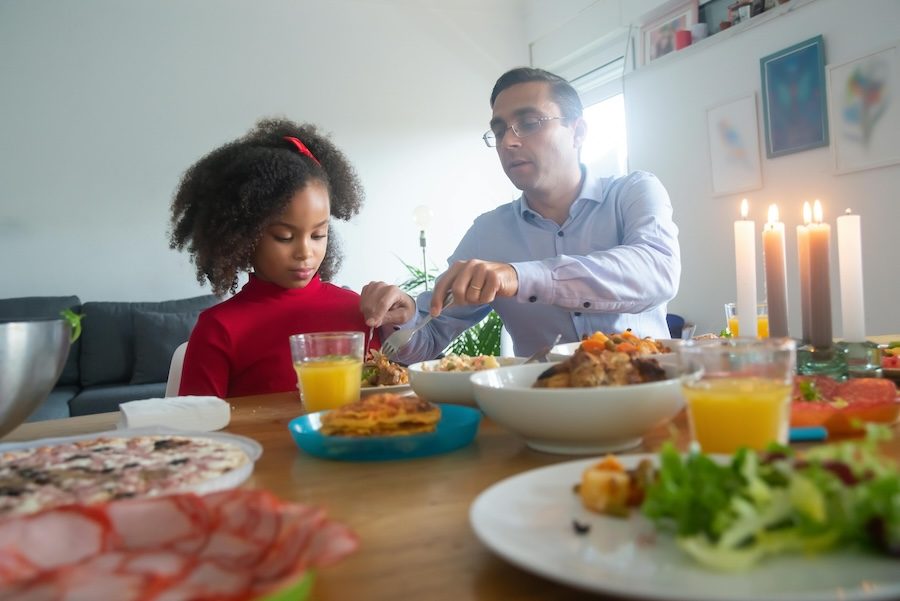 A white man in a button up shirt and glasses serves food to a Black girl with curly black hair, a red bow and red shirt. On the table there is a bowl of food, a glass of orange juice and a plate.