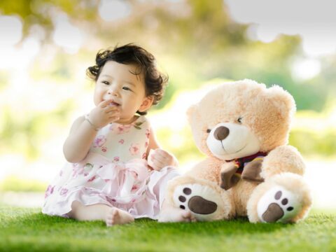 An East Asian baby with black hair has her right hand in her mouth. She is wearing a white and pink dress, sitting on the grass barefoot next to a light brown teddy bear that is almost the same size as her.