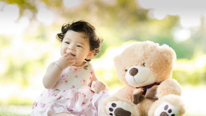 An East Asian baby with black hair has her right hand in her mouth. She is wearing a white and pink dress, sitting on the grass barefoot next to a light brown teddy bear that is almost the same size as her.