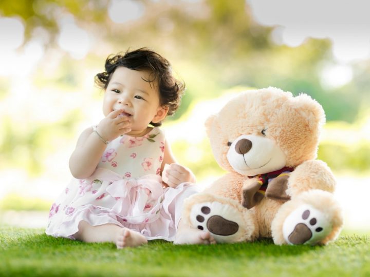 An East Asian baby with black hair has her right hand in her mouth. She is wearing a white and pink dress, sitting on the grass barefoot next to a light brown teddy bear that is almost the same size as her.