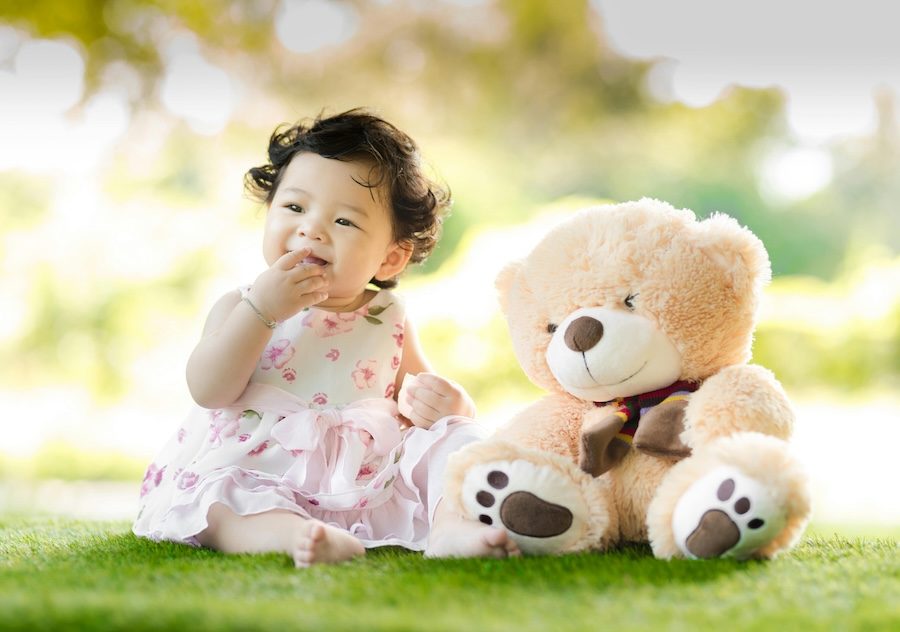 An East Asian baby with black hair has her right hand in her mouth. She is wearing a white and pink dress, sitting on the grass barefoot next to a light brown teddy bear that is almost the same size as her.