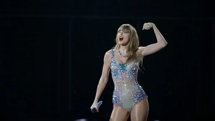 A white blond woman flexes her left arm and holds a mic in her right hand. She is wearing a short sparkly body suit.