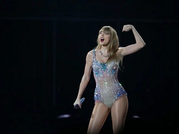 A white blond woman flexes her left arm and holds a mic in her right hand. She is wearing a short sparkly body suit.