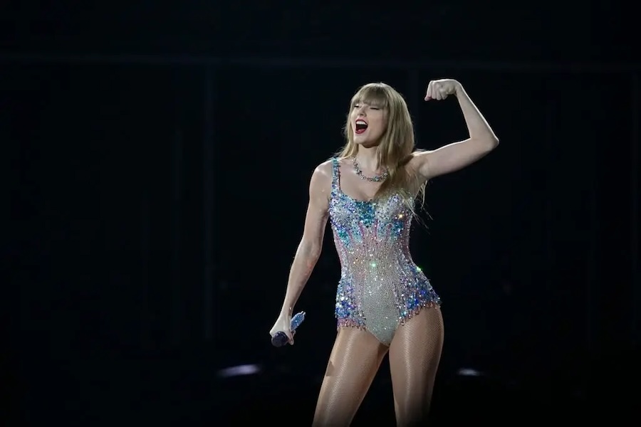 A white blond woman flexes her left arm and holds a mic in her right hand. She is wearing a short sparkly body suit.