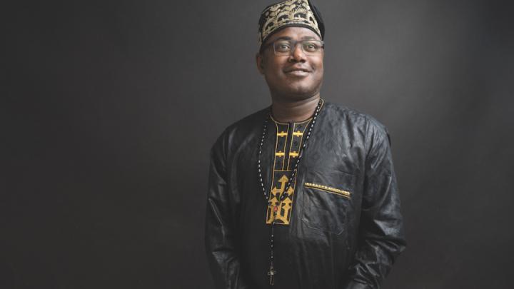 A Black man with glasses is dressed in traditional Nigerian clothing. He has a rosary hanging from his neck