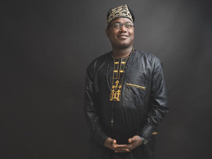 A Black man with glasses is dressed in traditional Nigerian clothing. He has a rosary hanging from his neck