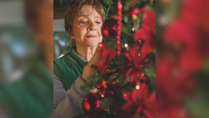 An older white woman with short brown hair places an ornament on a green Christmas tree with red decorations.