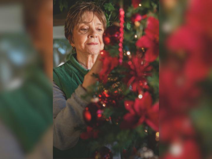 An older white woman with short brown hair places an ornament on a green Christmas tree with red decorations.