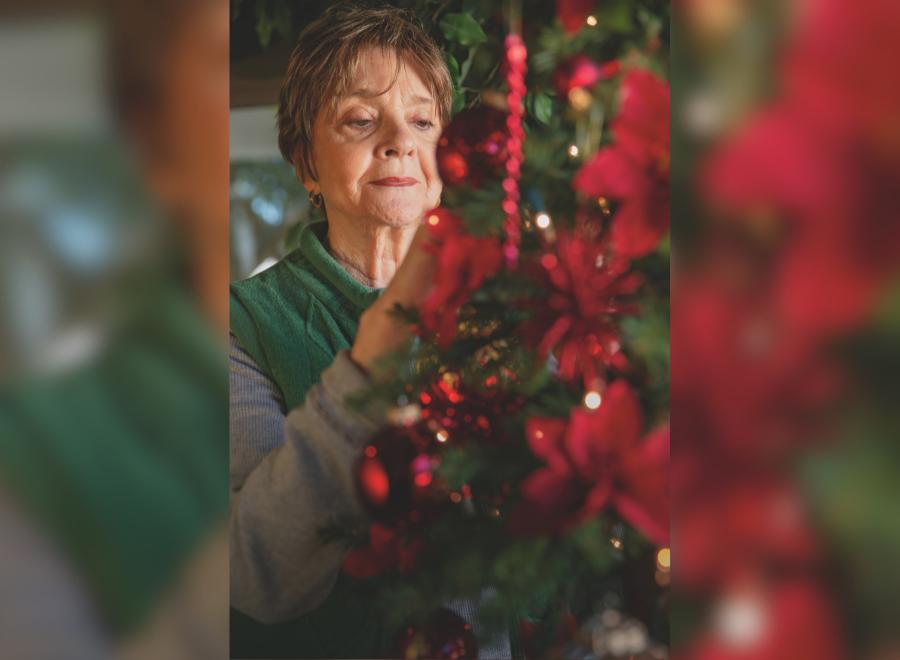 An older white woman with short brown hair places an ornament on a green Christmas tree with red decorations.