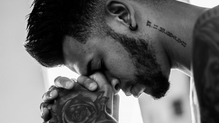 Black and white image of a man with a beard, a neck tattoo and tattoos on his hands praying