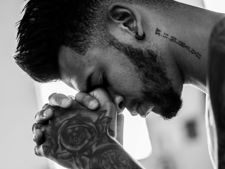 Black and white image of a man with a beard, a neck tattoo and tattoos on his hands praying