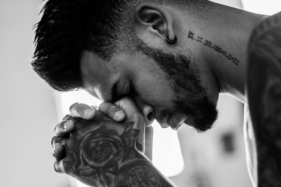 Black and white image of a man with a beard, a neck tattoo and tattoos on his hands praying