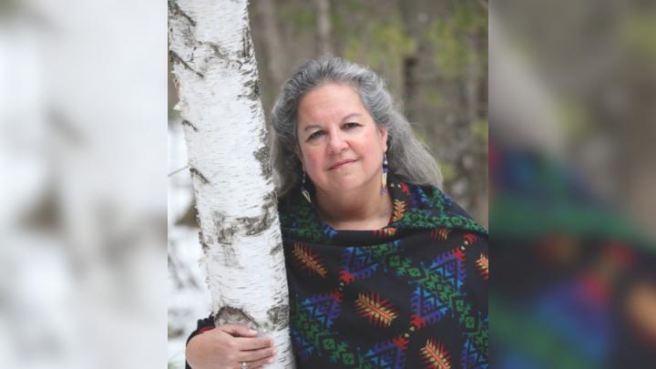 An Indigenous woman with grey hair stands with her right hand around a birch bark tree. She is wearing a blue and black knit wrap.