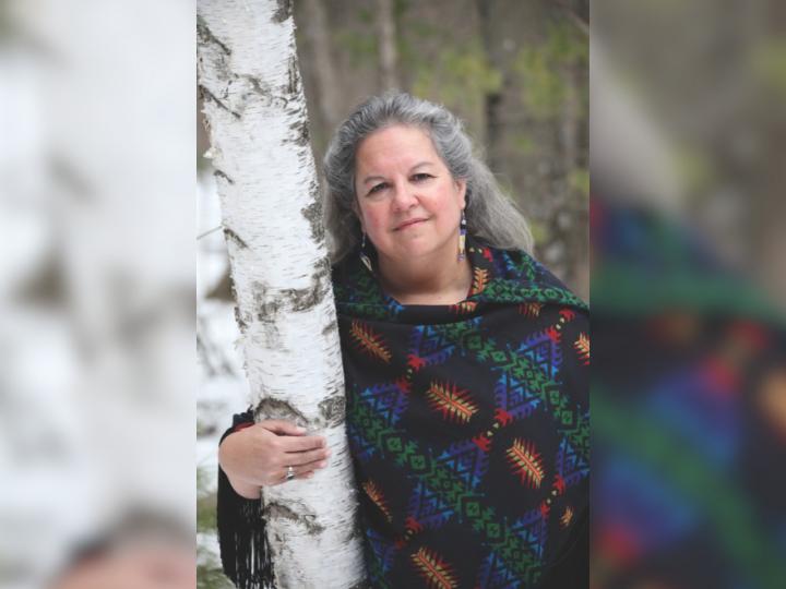 An Indigenous woman with grey hair stands with her right hand around a birch bark tree. She is wearing a blue and black knit wrap.