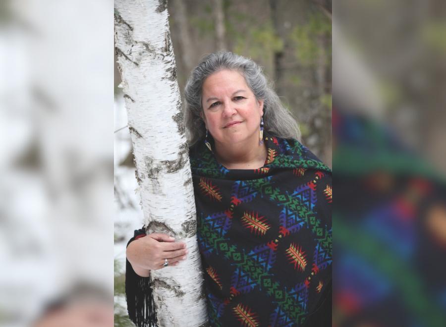 An Indigenous woman with grey hair stands with her right hand around a birch bark tree. She is wearing a blue and black knit wrap.