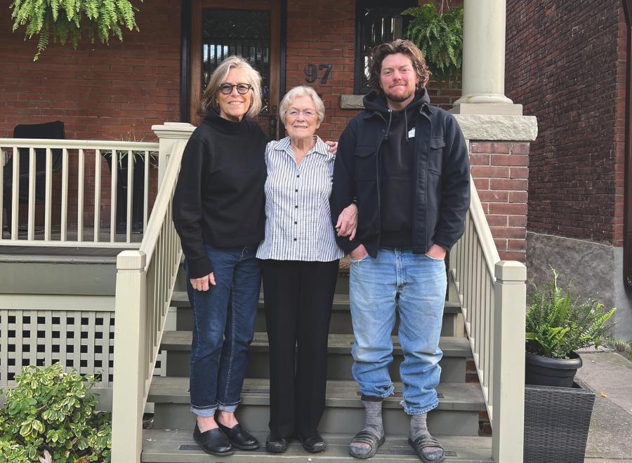 A middle aged white woman with short grey hair, in a black cardigan and blue jeans stands next to an older woman, who is a senior citizen. She is wearing a striped white and black button up, black slacks and black shoes. Her arm is linked with a young white man who has long brown hair, is wearing a black hoodie and blue jeans. They are standing on steps in front of a house.