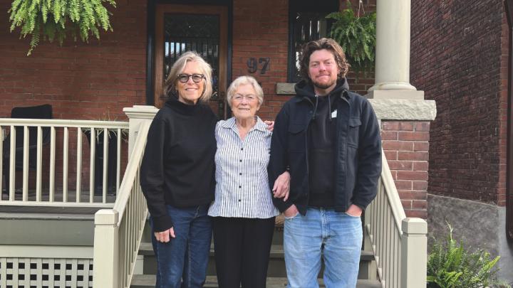 A middle aged white woman with short grey hair, in a black cardigan and blue jeans stands next to an older woman, who is a senior citizen. She is wearing a striped white and black button up, black slacks and black shoes. Her arm is linked with a young white man who has long brown hair, is wearing a black hoodie and blue jeans. They are standing on steps in front of a house.