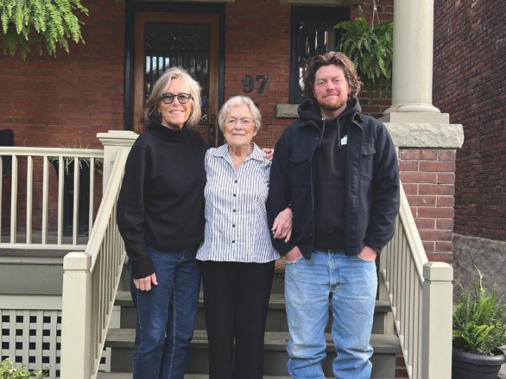 A middle aged white woman with short grey hair, in a black cardigan and blue jeans stands next to an older woman, who is a senior citizen. She is wearing a striped white and black button up, black slacks and black shoes. Her arm is linked with a young white man who has long brown hair, is wearing a black hoodie and blue jeans. They are standing on steps in front of a house.