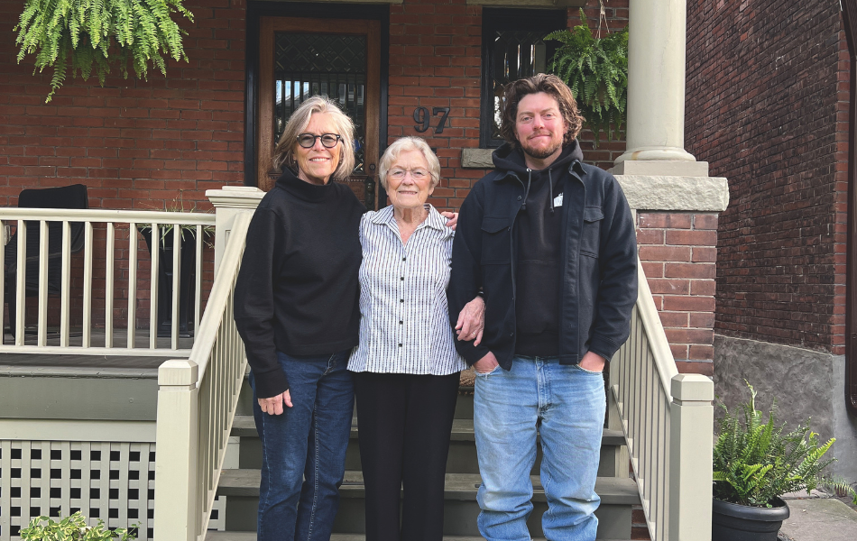 A middle aged white woman with short grey hair, in a black cardigan and blue jeans stands next to an older woman, who is a senior citizen. She is wearing a striped white and black button up, black slacks and black shoes. Her arm is linked with a young white man who has long brown hair, is wearing a black hoodie and blue jeans. They are standing on steps in front of a house.