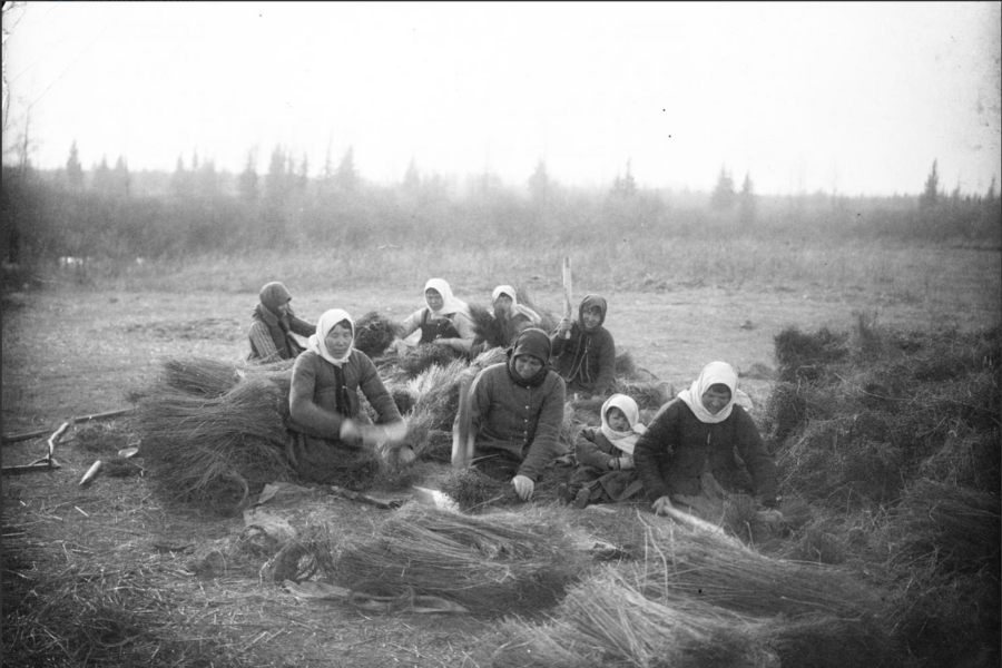 Black and white image of a group of women in headscarves sitting on the ground threshing flax in teams