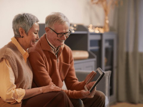 An older couple sit together looking at a tablet. They both have short grey hair. The man is wearing glasses and holding the tablet in his hands.