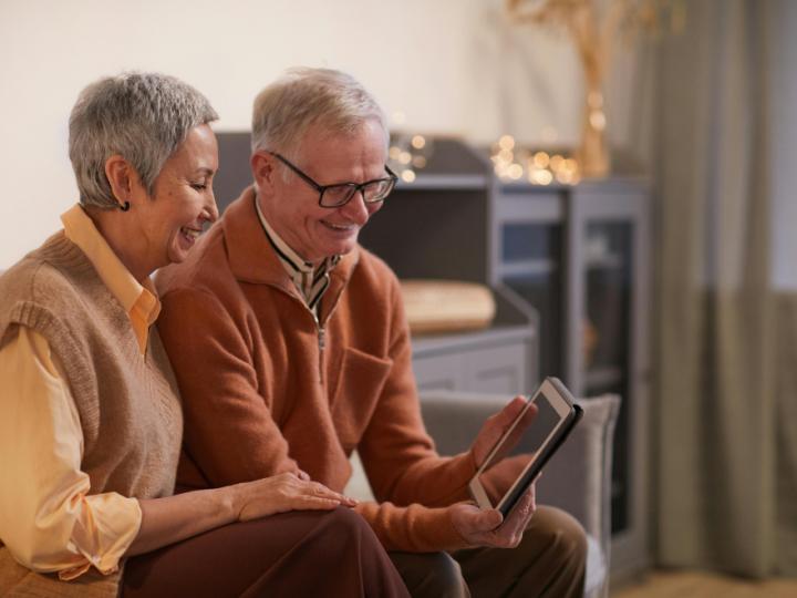 An older couple sit together looking at a tablet. They both have short grey hair. The man is wearing glasses and holding the tablet in his hands.